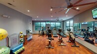 This single-floor fitness center offers a comprehensive workout environment designed for a wide range of exercise routines. The space features rich hardwood flooring throughout, lending warmth to the otherwise modern, clean aesthetic defined by gray walls and recessed lighting. Large black-framed windows line the perimeter, flooding the interior with natural light and providing open views of the surrounding outdoor scenery.

The cardio zone is generously equipped with multiple treadmills, elliptical trainers, and stationary cycling bikes, including spin-style bikes positioned in front of full-wall mirrors for form feedback. Wall-mounted flat-screen televisions are strategically placed throughout, ensuring entertainment during workouts. Ceiling fans complement the ventilation, contributing to a comfortable exercise environment.

The strength and functional training area features a cable resistance machine, a weight bench, a rack of dumbbells in varying sizes, and an assortment of stability and medicine balls. A bold geometric accent wall in navy, gray, and red adds a dynamic visual element to this zone.

Additional amenities include a water fountain station. The space benefits from a layout that allows for smooth circulation between equipment zones, making it practical and user-friendly for a variety of fitness goals.