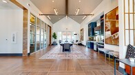 This single-story secondary clubhouse in Denton, TX is a well-appointed amenity facility offering a thoughtfully designed layout comprising a main gathering area, a hallway, an exercise room, and an office. The centerpiece of the clubhouse is the expansive main room, distinguished by soaring vaulted ceilings that create an airy, open atmosphere. Large-format wood-look tile flooring extends throughout, anchored by a styled area rug beneath a central dining-style table and chairs. A bold industrial-inspired pendant light fixture runs the length of the ceiling, complementing the built-in kitchenette featuring deep navy cabinetry, a mirrored tile backsplash, and under-counter appliances. Floor-to-ceiling glass partition walls allow natural light to flow freely, while exterior-facing doors provide outdoor views and additional brightness. A dedicated conference area off the hallway offers a formal meeting table surrounded by upholstered seating and a textured decorative wall panel. The exercise room is generously sized, carpeted in a plaid pattern, and outfitted with cardio machines, strength equipment, free weights, and full-length mirrors, with ceiling fans providing added comfort. A compact private office features a round worktable, rolling chairs, and a wall-mounted display screen, ideal for focused work sessions.