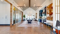 This single-story secondary clubhouse in Denton, TX is a well-appointed amenity facility offering a thoughtfully designed layout comprising a main gathering area, a hallway, an exercise room, and an office. The centerpiece of the clubhouse is the expansive main room, distinguished by soaring vaulted ceilings that create an airy, open atmosphere. Large-format wood-look tile flooring extends throughout, anchored by a styled area rug beneath a central dining-style table and chairs. A bold industrial-inspired pendant light fixture runs the length of the ceiling, complementing the built-in kitchenette featuring deep navy cabinetry, a mirrored tile backsplash, and under-counter appliances. Floor-to-ceiling glass partition walls allow natural light to flow freely, while exterior-facing doors provide outdoor views and additional brightness. A dedicated conference area off the hallway offers a formal meeting table surrounded by upholstered seating and a textured decorative wall panel. The exercise room is generously sized, carpeted in a plaid pattern, and outfitted with cardio machines, strength equipment, free weights, and full-length mirrors, with ceiling fans providing added comfort. A compact private office features a round worktable, rolling chairs, and a wall-mounted display screen, ideal for focused work sessions.