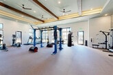 Located at 75 Caldwell Dr in Mount Holly, NC, this fitness center delivers a comprehensive workout environment within a generously sized, single-floor layout. The space is defined by soaring ceilings adorned with exposed wood beams and a coffered tray ceiling section, lending a warm, upscale aesthetic to the functional setting. Multiple ceiling fans and recessed lighting ensure the space remains well-ventilated and brightly lit throughout.

Large, black-framed windows line the walls, flooding the interior with abundant natural light and offering open views of the surrounding exterior. The flooring throughout is a durable, neutral-toned carpet suited for high-traffic fitness use.

The equipment selection is extensive and varied. A dedicated cardio zone features a row of modern treadmills and rowing machines positioned along the window walls. A central functional training rig in bold blue steel supports TRX suspension trainers and cable attachments, while a heavy boxing bag adds a combat fitness element. Strength training is well-represented with a full dumbbell rack, sled pull equipment, and plate-loaded implements.

A striking stacked-stone accent wall anchors one end of the space, complemented by decorative wood wall art and a wall-mounted television. Towel service and a storage station are conveniently positioned near the entrance, rounding out this polished and fully appointed fitness facility.