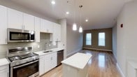 This single-floor space showcases a thoughtfully designed open-concept layout that seamlessly connects the kitchen and living area. The kitchen is outfitted with crisp white shaker-style cabinetry, white quartz countertops, and a classic grey subway tile backsplash that adds texture without overpowering the clean aesthetic. Stainless steel appliances, including a gas range, built-in microwave, and dishwasher, provide a polished, functional workspace. A central kitchen island with a white countertop offers additional prep space and serves as a natural dividing point between the kitchen and the adjoining living area. Pendant lights with cylindrical shades hang above, providing warm, focused illumination. The living area benefits from natural light streaming through multiple tall windows, enhancing the airy, open feel of the space. Light hardwood flooring runs continuously throughout, unifying the kitchen and living zones while adding warmth to the neutral grey and white color palette. Recessed lighting supplements the natural light, ensuring the space remains bright and well-lit at all times.