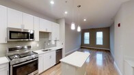 This single-floor space showcases a thoughtfully designed open-concept layout that seamlessly connects the kitchen and living area. The kitchen is outfitted with crisp white shaker-style cabinetry, white quartz countertops, and a classic grey subway tile backsplash that adds texture without overpowering the clean aesthetic. Stainless steel appliances, including a gas range, built-in microwave, and dishwasher, provide a polished, functional workspace. A central kitchen island with a white countertop offers additional prep space and serves as a natural dividing point between the kitchen and the adjoining living area. Pendant lights with cylindrical shades hang above, providing warm, focused illumination. The living area benefits from natural light streaming through multiple tall windows, enhancing the airy, open feel of the space. Light hardwood flooring runs continuously throughout, unifying the kitchen and living zones while adding warmth to the neutral grey and white color palette. Recessed lighting supplements the natural light, ensuring the space remains bright and well-lit at all times.