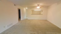 This single-floor space features an open and functional layout centered around a spacious living area. The room is finished with neutral-toned wall-to-wall carpeting that extends throughout, contributing to a warm and understated atmosphere. A ceiling fan with an integrated light fixture is centrally positioned overhead, providing both ventilation and ambient lighting to the main living space.

A defining architectural feature is the pass-through window connecting the living area to the kitchen, framed with a countertop ledge that allows for convenient interaction between the two spaces. This opening also allows light from the kitchen to filter into the living area, enhancing the overall brightness of the space. The kitchen itself appears to include standard cabinetry and overhead lighting visible through the pass-through.

White walls and standard baseboards give the interior a clean, simple aesthetic. A hallway extends from the main living area, leading to additional rooms beyond, including what appears to be a bathroom or bedroom visible in the background. A ceiling-mounted vent indicates a central HVAC system is in place. Overall, the space offers a straightforward, livable layout suited for everyday use.