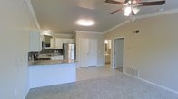 This single-floor space showcases an open-concept layout that combines the kitchen and living area into one cohesive, functional environment. The kitchen is well-appointed with white cabinetry, granite countertops, a stainless steel refrigerator, and a built-in microwave. A peninsula with a granite surface extends into the space, providing additional prep area and casual seating potential. The flooring transitions from light tile in the kitchen to neutral carpet in the living area, clearly defining each zone while maintaining a unified aesthetic. Crown molding runs along the upper walls, adding a refined architectural detail throughout. A ceiling fan with a light fixture is centrally positioned in the living area, promoting both airflow and illumination. Recessed and flush-mount lighting supplement the natural light filtering through the space. Doorways visible from the main area lead to additional rooms, including what appears to be a bathroom, offering convenient accessibility. The overall ambiance is bright, clean, and well-maintained, with a neutral color palette that lends itself to versatile interior styling.