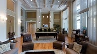 This sophisticated single-floor amenity space exudes a refined, upscale atmosphere designed for both relaxation and recreation. The room immediately draws the eye upward to its striking coffered ceiling, finished in crisp white with recessed lighting and elegant pendant fixtures that cast a warm, inviting glow throughout. The walls are dressed in a rich, earthy taupe palette with decorative panel molding, lending the space a classic, architectural character.

At the heart of the room sits a full-sized billiards table, serving as the focal point for recreation and social gatherings. Surrounding it are comfortable, dark brown leather armchairs and ottomans arranged over a large ornamental area rug, creating a welcoming lounge environment. A wall-mounted flat-screen television is positioned centrally for easy viewing from multiple seating areas.

Toward the rear of the space, a dining or gaming table with high-back chairs offers an additional area for group activities. Tall, floor-to-ceiling windows draped with sheer white curtains line one side of the room, bathing the interior in abundant natural light while providing a sense of openness. Lush indoor plants add a refreshing, organic touch to the overall design. Warm hardwood flooring throughout ties the space together, balancing the grandeur of the tall ceilings with an approachable, lived-in comfort.