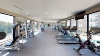 This fitness center is a spacious, single-floor facility designed to support a comprehensive workout experience. The space is divided into distinct functional zones, each outfitted with professional-grade equipment. The cardio area features a row of treadmills, elliptical trainers, and stationary bikes, all positioned near large windows that frame views of the surrounding outdoor areas and greenery, providing an open, energizing atmosphere during workouts. A mounted flat-screen television enhances the cardio experience.

The strength training section includes multiple cable and weight machines, along with a full dumbbell rack, offering a thorough range of resistance options. A rowing machine and additional specialty equipment round out the fitness offerings. Mirrored walls run along key sections of the gym, amplifying the sense of space and supporting form-checking during exercise.

Natural light floods the interior through floor-to-ceiling windows and large glass panels, connecting the interior to exterior courtyard views. Recessed ceiling lighting supplements natural light throughout. The flooring consists of durable, low-pile grey carpet suited for fitness use. The overall aesthetic is clean and modern, with neutral wall tones and a well-organized layout that maximizes usability and comfort.