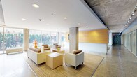 This thoughtfully designed single-floor amenity space combines industrial and contemporary elements to create a welcoming and functional environment. The lobby lounge is the centerpiece of the space, characterized by its exposed concrete ceiling and structural columns that lend an industrial character, while crisp white walls and recessed lighting maintain a bright, polished atmosphere. Polished terrazzo flooring runs throughout much of the space, transitioning seamlessly into carpeted seating areas that define distinct lounge zones.

Floor-to-ceiling glazing lines the perimeter, flooding the interior with abundant natural light and offering open views of the surrounding greenery and streetscape. Comfortable seating arrangements, featuring neutral-toned sofas and armchairs in cream and brown, are grouped around low cube tables and accented with colorful decorative objects and fresh floral arrangements. A warm wood-paneled feature wall anchors the main lounge area and is complemented by a mounted flat-screen display.

An adjacent area extends the lounge with additional seating and direct access to an outdoor entry. The space also includes a dedicated laundry facility, enclosed behind full-height glass partitions and equipped with rows of stacked washers and dryers. Throughout, the interplay of raw concrete, warm wood tones, and polished floors creates a balanced, contemporary ambiance suited for both relaxation and everyday convenience.