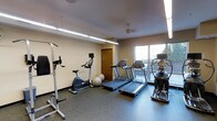 This fitness center is a dedicated exercise space designed to support a well-rounded cardio workout routine. The room features durable rubber flooring, ideal for high-traffic athletic use, and is illuminated by a combination of overhead fluorescent strip lighting and a ceiling fan that helps maintain air circulation throughout sessions. A large window along one wall floods the space with natural light and offers an outdoor view, creating an open and energizing atmosphere.

The cardio equipment lineup is the centerpiece of this gym, featuring multiple treadmills and an upright stationary bike from Matrix, catering to a variety of fitness preferences. An elliptical or rowing-style machine rounds out the cardio options on the far end of the room. Stability balls are neatly stored along the wall, adding a versatile functional training element to the space. A multi-use weight or resistance tower stands prominently near the entrance, offering additional strength training capability.

A warm-toned wooden door provides access to an adjoining area, contributing a subtle contrast to the neutral cream-colored walls. The overall layout is clean, organized, and efficiently arranged to maximize usable floor space while keeping all equipment easily accessible.