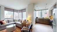 This contemporary high-rise unit offers a well-designed, single-floor layout with a cohesive modern aesthetic throughout. The open-concept living area is bathed in natural light from floor-to-ceiling windows, framing sweeping urban skyline views. The space is anchored by a generous sectional sofa, complemented by warm-toned area rugs and light hardwood flooring that extends throughout the unit.

Adjacent to the living area, a compact yet functional kitchenette features dark cabinetry, a sink, and a coffee station — ideal for everyday convenience. The full kitchen is outfitted with sleek gray cabinetry, quartz countertops, a smooth-top electric range, built-in microwave, and stainless steel Bosch appliances, offering a polished and highly functional cooking space.

The bedroom is a serene retreat, featuring large windows that frame city views, light hardwood floors, clean white walls, and ample closet space. The room's neutral palette creates a calm and restful atmosphere.

The bathroom is finished with large-format dark floor tiles, a floating white vanity with an illuminated LED mirror, and a full shower with contrasting tile accents — delivering a clean, spa-like feel.

Throughout the unit, the design emphasizes clean lines, neutral tones, and thoughtful finishes, creating a cohesive and sophisticated urban living environment.