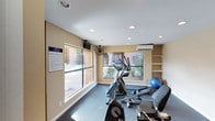 This single-level fitness center offers a practical and welcoming environment for residents or guests seeking a dedicated space for exercise. The room is outfitted with cardio equipment, including an elliptical trainer and stationary bike, as well as a stability ball and additional workout accessories, providing a range of options for varied fitness routines. Large windows along the walls flood the space with natural light and offer views of the surrounding outdoor courtyard and landscaping, creating an open and energizing atmosphere. The flooring features durable, dark carpeting suited for gym use, while recessed lighting in the textured ceiling ensures the space remains well-lit throughout. A wall-mounted air conditioning unit provides climate control for year-round comfort, complemented by a ceiling-mounted fan for additional airflow. Built-in shelving along one wall offers convenient storage for equipment and accessories. A posted fitness center rules placard is prominently displayed near the entrance, underscoring the organized and community-oriented nature of the facility. The warm, neutral-toned walls contribute to a clean and functional aesthetic throughout the space.