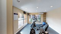 This single-level fitness center offers a practical and welcoming environment for residents or guests seeking a dedicated space for exercise. The room is outfitted with cardio equipment, including an elliptical trainer and stationary bike, as well as a stability ball and additional workout accessories, providing a range of options for varied fitness routines. Large windows along the walls flood the space with natural light and offer views of the surrounding outdoor courtyard and landscaping, creating an open and energizing atmosphere. The flooring features durable, dark carpeting suited for gym use, while recessed lighting in the textured ceiling ensures the space remains well-lit throughout. A wall-mounted air conditioning unit provides climate control for year-round comfort, complemented by a ceiling-mounted fan for additional airflow. Built-in shelving along one wall offers convenient storage for equipment and accessories. A posted fitness center rules placard is prominently displayed near the entrance, underscoring the organized and community-oriented nature of the facility. The warm, neutral-toned walls contribute to a clean and functional aesthetic throughout the space.