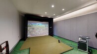 Located at 7971 S Sixth St in Oak Creek, Wisconsin, this dedicated indoor golf simulator bay delivers a fully immersive golf experience in a compact, purpose-built space. The room is finished with wall-to-wall artificial turf flooring, providing an authentic feel underfoot across both the hitting area and the surrounding floor. A premium Foresight Sports simulator system serves as the centerpiece, projecting vivid, detailed course imagery onto a large impact screen framed by a black surround wall, creating a focused and distraction-free hitting environment. The simulator station includes a dedicated equipment cart housing the launch monitor and projector setup, positioned to precisely track every shot. Along the side wall, upholstered acoustic panels in a neutral gray tone line the perimeter, contributing to sound dampening and a clean, professional aesthetic. Recessed can lighting is arranged across a tray ceiling, supplemented by soft indirect lighting that evenly illuminates the space without glare. A dedicated hitting mat sits atop the turf, and golf balls are readily accessible near the tee area. Seating and a small bar-height table are positioned along the side wall for guests or coaching sessions. The room also features HVAC vents for climate control and a private entry door, making it a self-contained, fully equipped simulator suite.