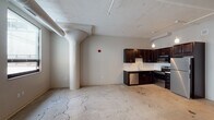 This single-floor unit showcases a distinctive industrial aesthetic rooted in its original architectural character. The open-concept living and kitchen area is defined by polished exposed concrete floors with natural veining, a textured ceiling with visible ductwork and track lighting, and a prominent rounded structural column that adds to the loft-like character of the space. Windows along one wall allow natural light to filter in, offering views of the surrounding building exterior.

The kitchen is well-appointed with rich dark espresso cabinetry, light stone countertops, a white subway tile backsplash, and a full suite of stainless steel appliances, including a refrigerator, electric range, built-in microwave, and dishwasher. Pendant lighting above the kitchen area provides warm task illumination.

The bedroom is a quieter, carpeted space with neutral wall tones, offering a softer contrast to the industrial main area. It connects to a clean, functional bathroom featuring hexagonal dark tile flooring, a white vanity with sink, a full tub-shower combination, and a wall-mounted mirror with overhead lighting. The overall ambiance is a balanced blend of raw, urban character and modern residential finishes.