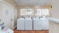 This shared laundry room is a functional and well-maintained facility designed to accommodate multiple users efficiently. The space features warm beige walls and rich hardwood flooring that lend a clean, welcoming atmosphere. Multiple top-loading washing machines and dryers are arranged in a row along the walls, providing ample capacity for residents. A built-in shelving ledge above the machines offers convenient space for laundry supplies and personal items. Large windows fitted with vertical blinds line the walls, allowing generous natural light to fill the room and providing views of the surrounding outdoor greenery. The vaulted ceiling with exposed white beams adds a sense of openness and airiness to the space. Wall-mounted light fixtures supplement natural lighting for evening use. A fire extinguisher is mounted on the wall, reflecting a commitment to safety. The room also includes a dedicated folding counter surface, making laundry tasks more convenient. Access is provided through a solid door, and the overall layout is thoughtfully organized to ensure ease of movement throughout the space. The combination of practical amenities, bright natural light, and clean finishes makes this laundry room a comfortable and efficient facility.