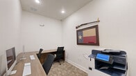 Located at 562 Centennial Pkwy in Tyler, Texas, this single-room office space offers a clean, efficient environment designed for productive work. The room features neutral white walls and patterned carpet tiles in warm beige tones that lend a professional yet welcoming atmosphere throughout.

The workspace is anchored by an L-shaped built-in wooden desk along one wall, providing ample surface area for multiple workstations. Two desktop computers are set up side by side, accompanied by ergonomic task chairs, making the space well-suited for collaborative or dual-user work. An additional desk section extends perpendicularly, offering further workspace flexibility.

A dedicated area houses a full-sized multifunction printer and copier, keeping essential office equipment organized and accessible. Prominent double doors with a clean white panel design provide both visual appeal and practical storage or access to an adjoining area. The doorway opens to a hallway with hardwood-style flooring, connecting the office to the broader facility.

A decorative Texas flag painting and a mounted rifle display on the wall add a distinctive regional character to the space. Ceiling-mounted vents ensure proper climate control, while the overall layout maximizes utility within a compact footprint. This office is a streamlined, functional workspace tailored for day-to-day professional operations.