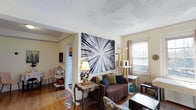 This single-floor space offers a warm and inviting atmosphere highlighted by rich hardwood flooring that runs throughout the main living area. The living room serves as the central gathering space, featuring a large, eye-catching black-and-white wall art piece depicting an abstract tree canopy, which anchors the room with a bold artistic statement. A comfortable sofa dressed in earthy tones — including deep brown, olive green, and dark accent pillows — provides a relaxed seating arrangement. A classic wooden side table with a decorative table lamp adds a charming, eclectic touch to the space. Natural light filters in through a window, brightening the room and creating an airy feel. A textured ceiling adds architectural character overhead. The living area opens directly into a compact kitchen, visible through a doorway and equipped with standard appliances, including a refrigerator. The layout flows naturally between spaces, making the overall footprint feel efficient and well-connected. Additional functional touches, such as a multi-tiered cat tree near the window, reflect a lived-in, personalized environment. The overall ambiance is casual, creative, and comfortable, with a mix of decorative elements that give the space a distinctive, homey character.