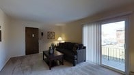 This single-floor residential unit offers a comfortable and functional layout designed for everyday living. The living room serves as the centerpiece of the space, featuring plush wall-to-wall carpeting that adds warmth underfoot. A large sliding glass door dressed with vertical blinds dominates one wall, allowing an abundance of natural light to fill the room while offering direct access to an outdoor area and views beyond. The living area comfortably accommodates a sectional sofa and a dark wood coffee table, creating a welcoming and well-appointed gathering space. Warm ambient lighting from a table lamp complements the natural light, contributing to the room's inviting atmosphere. Neutral-toned walls and a smooth white ceiling keep the space feeling bright and open. A solid wood entry door provides a sense of privacy and security at the entrance. Subtle decorative touches, including wall art and accent pieces, add personality without overwhelming the clean, understated aesthetic. The overall ambiance is calm and residential, with a practical design that balances comfort and simplicity throughout.
