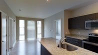This single-floor residence offers a thoughtfully designed open-concept layout that seamlessly connects the kitchen and living areas. The kitchen serves as the centerpiece of the space, showcasing a large island with striking granite countertops, an undermount sink with a brushed nickel faucet, and sleek dark cabinetry that provides ample storage. Track lighting above the kitchen island adds focused illumination, complementing the recessed lighting throughout. The living area benefits from generous natural light streaming through multiple large windows dressed with horizontal blinds, creating a bright and welcoming atmosphere. Light hardwood-style flooring runs throughout, adding warmth and continuity to the open floor plan. Neutral gray walls provide a clean, contemporary backdrop that suits a variety of design preferences. A closet with built-in shelving offers convenient storage, while an adjoining room — visible through a clean white-trimmed doorway — adds further functional space. The overall design aesthetic is modern and understated, with quality finishes and a well-balanced combination of natural and artificial lighting that makes the space feel both practical and inviting.