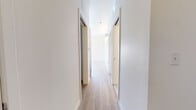 This single-floor unit showcases a thoughtfully designed layout with a consistent modern aesthetic carried throughout each space. Light wood-tone flooring runs continuously across the main living areas and bedrooms, lending warmth and visual cohesion to the interior.

The kitchen is a standout feature, equipped with a full suite of stainless steel appliances including a refrigerator, electric range with overhead microwave, and dishwasher. Textured wood-grain cabinetry in warm taupe tones extends from lower cabinets to upper storage, maximizing functionality. White countertops provide a clean contrast, while large windows flood the space with natural light and offer outward views.

The living area benefits from notably high ceilings, enhancing the sense of openness. A sliding glass door allows access to the exterior, further brightening the room. Built-in linear wall lighting in the bedroom areas adds a sleek architectural detail.

The hallway connects rooms efficiently with clean white walls and matching wood flooring. The bathroom features large-format light stone-effect floor tiles, white subway tile in the shower area, a wood-tone vanity with a white countertop, and simple chrome fixtures.

Overall, the unit presents a clean, minimalist character defined by neutral tones, quality finishes, and an abundance of natural light.