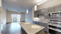 Located at ArtHaus 2828 in Dallas's vibrant 75204 corridor, this one-bedroom, one-bathroom residence offers a thoughtfully designed open-concept layout. The combined kitchen and living area serves as the heart of the home, anchored by a generous granite island with pendant lighting overhead. The kitchen is outfitted with sleek grey cabinetry, a striking stone tile backsplash, stainless steel appliances including a double oven range, built-in microwave, and a French door refrigerator. Rich dark hardwood flooring extends throughout the main living area, complementing the modern, contemporary aesthetic. Sliding glass doors open to a private balcony with cable railing, filling the living space with natural light. The bedroom features plush carpet, a ceiling fan, and a large window with city views. Three closets, including a well-organized walk-in with built-in shelving and dual hanging rods, provide ample storage. The bathroom offers a double-sink granite vanity with grey cabinetry, a wide framed mirror, and clean large-format tile flooring. A dedicated laundry area and utility room round out this efficiently designed urban residence.