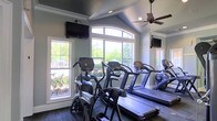 Located at 400 Stone Lion Dr in Durham, North Carolina, this fitness center offers a well-appointed workout environment designed for both comfort and functionality. The space features vaulted ceilings with crown molding, giving the room an open and airy feel. Large arched and rectangular windows flood the interior with abundant natural light while providing pleasant views of the surrounding landscaped greenery. A ceiling fan overhead ensures comfortable air circulation during workouts. The cardio equipment area is outfitted with Cybex treadmills, elliptical cross-trainers, and a stair climber, all arranged to take advantage of the bright, window-facing layout. Wall-mounted flat-screen televisions are positioned for easy viewing from multiple workout stations, enhancing the overall exercise experience. Full-length mirrors line one wall, visually expanding the space and allowing users to monitor their form. The neutral color palette of soft blue-gray walls paired with light trim creates a clean and motivating atmosphere. The combination of thoughtful design, quality equipment, and ample natural light makes this a functional and inviting fitness facility.