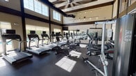 Located at 130 S Dixie Way in Roseland, Indiana, this fitness center is a standout facility designed to support a comprehensive workout experience. The space is defined by its soaring vaulted ceilings adorned with dark-stained exposed wood beams, creating a dramatic architectural character that sets it apart from a typical gym environment. Large, multi-pane windows line the walls from floor to upper clerestory level, flooding the interior with abundant natural light and offering views of an outdoor pool area, adding a refreshing sense of openness to every workout. The durable rubber flooring provides both safety and comfort throughout the space. The cardio zone is generously appointed with multiple treadmills, elliptical trainers, and stationary bikes from Life Fitness, each equipped with individual display screens for entertainment and performance tracking. Wall-mounted flat-screen televisions are positioned throughout for additional viewing. The strength training area features a multi-station cable machine and a full rack of dumbbells, complemented by floor-to-ceiling mirrors that visually expand the space and support proper form. Overhead, large ceiling fans and track lighting ensure consistent airflow and visibility. The overall ambiance is bright, modern, and inviting, making this fitness center a well-rounded destination for residents seeking a high-quality exercise facility.