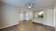 This single-floor residence showcases a thoughtfully designed open-concept layout that seamlessly connects the living and kitchen areas. The spacious living room features warm wood-look hardwood flooring, neutral-toned walls, and a ceiling fan with integrated lighting, creating a bright and comfortable atmosphere. Recessed track lighting in an adjoining nook adds versatility to the space, making it ideal for use as a dining or flex area.

The kitchen is accessible via a raised peninsula counter finished with granite countertops, offering both a functional workspace and a casual dining or seating option. White cabinetry extends to the upper walls, providing ample storage, while stainless steel appliances and a tasteful tile backsplash contribute to a clean, contemporary aesthetic. A built-in appliance such as a microwave or oven is also visible within the cabinetry wall.

The flooring flows consistently throughout, unifying the open space. HVAC vents are integrated discreetly into the walls and ceiling, ensuring year-round comfort. A doorway leads to additional rooms beyond the main living area, suggesting further private spaces. Overall, the residence presents a modern, well-maintained interior with a neutral palette that offers a versatile and welcoming environment.