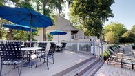 Pool 3601 is an outdoor deck space designed for leisure and relaxation, nestled among mature trees draped with Spanish moss that provide natural shade and a tranquil atmosphere. The expansive wooden deck serves as the central feature, offering ample open space for guests to unwind in a comfortable, resort-style environment.

The deck is furnished with wrought iron tables and chairs, complemented by large blue market umbrellas that provide additional shade during sunny days. Multiple seating arrangements are spread across the deck, accommodating both small groups and larger gatherings with ease.

A white picket-style railing borders the perimeter of the deck, defining the space while maintaining an open, airy feel. Steps lead down from the deck to a paved walkway, seamlessly connecting the area to the surrounding grounds. In the background, a neighboring building with a elevated balcony and white railing is visible, contributing to the cohesive aesthetic of the overall community.

The lush, green tree canopy overhead creates a naturally cool and inviting environment, making this pool deck an appealing outdoor retreat suited for casual lounging and social gatherings alike.