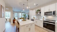This single-story, one-bedroom, one-bathroom unit is thoughtfully designed to maximize functionality within a compact footprint. The open-concept layout connects the kitchen and living area seamlessly, creating an airy, cohesive living space bathed in natural light from large windows fitted with blinds and sheer curtains.

The kitchen serves as a central highlight, featuring crisp white shaker cabinetry, a light grey subway tile backsplash, quartz countertops, and a full suite of stainless steel appliances including a range, dishwasher, microwave, and refrigerator. A sizeable central island with a quartz top and built-in storage provides bar seating for two and functions as an ideal casual dining and prep area. Elegant glass pendant lights hang above the island, adding a refined touch. Open shelving above the cabinetry offers additional display and storage space.

Light wood-tone flooring runs consistently throughout the unit, enhancing the bright, modern aesthetic. The bedroom accommodates a full-size bed comfortably and benefits from multiple windows that bring in abundant natural light. A dedicated walk-in closet with wire shelving provides practical storage. The bathroom features a white vanity with quartz countertop, undermount sink, a large mirror, and a tall storage cabinet, maintaining the clean, contemporary design language found throughout the unit.