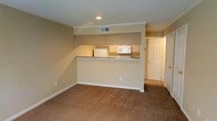 This two-floor residential space offers a practical and comfortable layout designed for everyday living. The main living area is finished with wall-to-wall carpeting in a warm beige tone, creating a cozy and inviting atmosphere. A defining architectural feature is the half-wall pass-through that connects the kitchen to the living area, providing an open feel while maintaining a clear distinction between spaces. The kitchen is equipped with white appliances, including a refrigerator and microwave, set against granite-look countertops and light-toned cabinetry. An overhead soffit above the kitchen adds a structured, built-in aesthetic to the space. Neutral beige walls are accented with crisp white baseboards and crown molding, lending a clean and cohesive look throughout. Recessed lighting in the ceiling provides focused illumination, complemented by a ceiling vent that ensures comfortable climate control. A paneled white door visible in the background leads to additional rooms or storage, hinting at further functional spaces within the home. The overall ambiance is understated and move-in ready, with a straightforward design that prioritizes utility and comfort across both floors.
