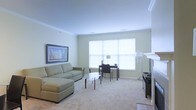 This well-appointed single-floor space offers a clean, functional layout designed for both relaxation and productivity. The living area is bathed in natural light streaming through a large multi-pane window dressed with white roller shades, creating a bright and airy atmosphere. Crown molding along the ceiling adds a refined architectural touch, while plush wall-to-wall carpeting lends warmth and comfort underfoot.

The living room is furnished with a spacious sage-green sectional sofa paired with a sleek glass coffee table, providing an inviting area to unwind. A framed landscape artwork adorns the soft-toned wall, adding a tasteful decorative accent. Adjacent to the seating area, a compact desk and chair arrangement makes excellent use of the natural light from the window, offering a dedicated workspace within the same open area.

A white fireplace mantle is visible along one wall, contributing an elegant focal point and classic charm to the room. The neutral, light-toned walls throughout enhance the sense of openness and allow for versatile interior styling. The overall ambiance of the space is calm, clean, and inviting — well-suited for comfortable everyday living.