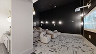 This sophisticated screening room delivers an immersive cinematic experience through a carefully curated design that balances drama and comfort. The room is defined by its striking matte black walls that stretch to impressive heights, creating a darkened, theater-like atmosphere ideal for viewing. A large projection screen anchors one end of the space, positioned for optimal sightlines from every seat. Ambient illumination is provided by a series of elegant gold teardrop pendant sconces mounted along the walls, casting a warm, intimate glow throughout the room. The seating arrangement features multiple rows of plush, cream-upholstered lounge chairs and sofas, each paired with rustic wood side tables that introduce a natural texture against the room's monochromatic palette. Underfoot, a bold black-and-white abstract patterned carpet adds graphic visual interest and ties the design together cohesively. The entryway transitions from dark tile flooring, providing a subtle delineation between the corridor and the main screening area. Wall-mounted speakers are integrated discreetly throughout, supporting a full audio experience. A piece of contemporary wall art near the entry adds a finishing artistic touch. Overall, the space achieves a refined, upscale aesthetic that is both functional and visually compelling.