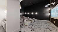 This sophisticated screening room delivers an immersive cinematic experience through a carefully curated design that balances drama and comfort. The room is defined by its striking matte black walls that stretch to impressive heights, creating a darkened, theater-like atmosphere ideal for viewing. A large projection screen anchors one end of the space, positioned for optimal sightlines from every seat. Ambient illumination is provided by a series of elegant gold teardrop pendant sconces mounted along the walls, casting a warm, intimate glow throughout the room. The seating arrangement features multiple rows of plush, cream-upholstered lounge chairs and sofas, each paired with rustic wood side tables that introduce a natural texture against the room's monochromatic palette. Underfoot, a bold black-and-white abstract patterned carpet adds graphic visual interest and ties the design together cohesively. The entryway transitions from dark tile flooring, providing a subtle delineation between the corridor and the main screening area. Wall-mounted speakers are integrated discreetly throughout, supporting a full audio experience. A piece of contemporary wall art near the entry adds a finishing artistic touch. Overall, the space achieves a refined, upscale aesthetic that is both functional and visually compelling.