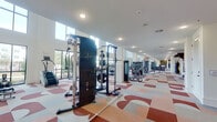 This well-appointed fitness center is designed to accommodate a wide range of workout needs within a generously proportioned, single-floor layout. The space is defined by its striking double-height ceilings, which create an open, airy atmosphere enhanced by rows of tall windows along the perimeter walls. These expansive windows flood the interior with natural light and offer views of the surrounding outdoor environment, contributing to an energizing workout atmosphere.

The flooring throughout the facility features a bold, patterned carpet in shades of red, gray, and white, adding a dynamic visual element to the space. Large ceiling fans are mounted overhead, ensuring consistent airflow across the workout areas. A mezzanine-level overlook with a railing is visible from the main floor, adding architectural depth to the interior.

Equipment is thoughtfully arranged throughout the space and includes treadmills, ellipticals, stair climbers, cable machines, an Axis functional trainer, and a full dumbbell rack accompanied by a weight bench. Full-length mirrors along one wall enhance the sense of space and allow users to monitor their form. A dedicated area with built-in shelving, a beverage refrigerator, and wall-mounted televisions provides added convenience. A staircase is accessible from within the facility.