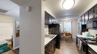 This single-floor residence offers a thoughtfully designed layout that balances functionality and comfort across its key living spaces. The galley-style kitchen is a standout feature, outfitted with dark espresso cabinetry, light countertops, black appliances including a full-size refrigerator and electric range, and a stainless steel sink. Upper and lower cabinets provide generous storage, and wood-look flooring ties the space together seamlessly. Adjacent to the kitchen, the dining area benefits from natural light streaming through a window dressed with white curtains and blinds, creating a bright and welcoming atmosphere for meals. A colorful wall accent adds a touch of personality to the space. The living room is generously sized, featuring the same wood-look flooring and neutral wall tones that lend a clean, modern feel throughout. The bedroom offers plush carpeting, a wide sliding-door closet for ample storage, and a window that allows natural light to filter in. The bathroom is efficiently laid out with a walk-in shower, toilet, and vanity, all finished with wood-look flooring and neutral tones. Textured ceilings are present throughout, contributing to the space's residential character.