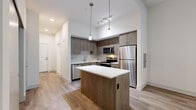 Located at Bear Creek Way NE in Redmond, Washington, this single-floor residence offers a well-designed layout with modern finishes throughout. The kitchen serves as a central highlight, showcasing warm-toned wood cabinetry paired with a geometric tile backsplash, crisp white quartz countertops, and a full suite of stainless steel appliances including a refrigerator, range, microwave, and dishwasher. A central island with a white countertop provides additional workspace and casual seating. Pendant lighting and a track light fixture illuminate the space effectively, complementing the recessed lighting overhead. Wood-style plank flooring runs consistently throughout the living areas, adding warmth and visual continuity. The living space is generously proportioned, with tall ceilings that enhance the sense of openness. Large windows fitted with horizontal blinds allow considerable natural light to fill the room, while a ceiling fan adds practical comfort. The bathroom features large-format marble-look tile flooring, a wood-toned vanity with white countertop, a wall-mounted cabinet for storage, and a tiled shower area. The overall design aesthetic is clean and contemporary, with a neutral palette of whites and warm wood tones creating a cohesive, inviting atmosphere throughout the unit.