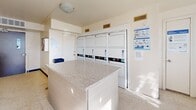 This shared laundry facility is a clean, efficiently organized space designed to serve a multi-unit building. The single-floor layout is divided into two connected areas that work together to provide a comprehensive laundry experience. The main area features a row of commercial-grade front-load washing machines lining one wall, while a bank of stacked dryer units occupies the opposite end, maximizing the available floor space. A central granite-topped folding island provides a dedicated surface for sorting and folding laundry, offering both functionality and a clean aesthetic. The secondary area includes a utility sink set beneath a window, framed by white tile walls and cabinetry below, allowing natural light to filter in and brighten the workspace. Throughout the facility, durable terrazzo flooring adds a classic, easy-to-maintain finish. The walls are painted in neutral white tones, contributing to a bright and hygienic atmosphere. Instructional signage is posted throughout for user guidance. Overhead flush-mount lighting ensures consistent illumination across the space. A payment station is conveniently located within the room, and the facility is accessible via two doorways. Overall, the space is thoughtfully laid out to accommodate multiple users simultaneously with ease and efficiency.
