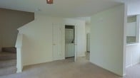 This two-story home offers a functional and straightforward layout across both floors. The image showcases a lower-level living area finished with wall-to-wall carpeting in a neutral tone, contributing to a clean and understated aesthetic. The walls are painted in a soft off-white, creating a bright and open feel throughout the space. A ceiling light fixture is visible overhead, providing ambient illumination to the room.

A notable feature of this level is the dedicated in-unit laundry closet, which houses a stacked washer and dryer unit — a practical convenience tucked neatly behind a door. Adjacent to the laundry closet is an additional door, likely leading to a storage or utility space. A hallway visible in the background connects to further areas of the home, where tile flooring transitions from the carpeted living space, and a refrigerator can be seen, indicating the kitchen is nearby.

The layout suggests a logical separation of living and utility functions on this floor. With its neutral finishes, in-unit laundry, and multi-floor configuration, this home offers a comfortable and practical living environment suited to everyday needs.