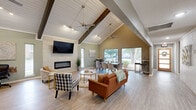 Located at 3549 Curry Ln in Abilene, TX, this single-story space offers a thoughtfully designed open-concept layout that maximizes both functionality and visual appeal. The centerpiece of the space is the expansive Great Room, defined by soaring vaulted ceilings with white shiplap paneling and striking dark wood exposed beams that draw the eye upward. Dual ceiling fans and track lighting provide both comfort and ambiance. A sleek linear electric fireplace set into a white tile accent wall serves as the room's focal point, flanked by a large-screen television above. Light wood-look flooring runs throughout, complementing the soft sage green and warm greige wall tones. Generous windows along the exterior walls flood the space with natural light and offer views of the surrounding greenery. The open layout accommodates distinct seating, dining, and workspace zones, all flowing seamlessly together. The Kitchen features gray shaker cabinetry topped with white countertops, stainless steel appliances including a range with a wall-mounted hood vent, open floating shelves, and a textured geometric tile backsplash that adds a modern, industrial touch. A pendant light illuminates the sink area, completing the kitchen's contemporary aesthetic.
