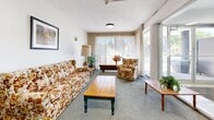 Located at 1791 King St in Hamilton, Ontario, this ground-floor common area serves as the main lobby and resident lounge for the building. The entrance corridor is defined by striking terrazzo flooring with inlaid tile detailing, leading visitors through a well-lit passageway framed by distinctive cylindrical columns clad in mosaic tile. Glass doors at the front entry allow natural light to filter into the space, creating a bright and inviting atmosphere upon arrival.

The lobby area is equipped with practical amenities, including a Canada Post parcel locker station and a building notice board, ensuring residents have convenient access to mail and building communications. The transition from the entrance corridor into the lounge area is seamless, where carpet flooring softens the space and adds warmth.

The lounge is a comfortable, generously proportioned sitting area furnished with upholstered seating, a coffee table, and decorative plants that contribute to a relaxed, welcoming ambiance. Large sliding glass doors and expansive windows flood the lounge with natural light and offer views of the surrounding greenery, enhancing the sense of openness. Textured ceilings and neutral white walls throughout maintain a clean, understated aesthetic, making this shared space both functional and comfortable for residents and guests alike.