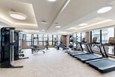 This well-equipped fitness center at 3100 Lake Villa Dr in Metairie, Louisiana offers a spacious, single-floor layout designed to support a comprehensive workout experience. The room is defined by its modern architectural design, featuring a coffered ceiling with layered tray details, ambient cove lighting, and a series of flush circular light fixtures that create a clean, polished aesthetic throughout.

Large floor-to-ceiling windows line the perimeter walls, flooding the space with abundant natural light and providing open views of the surrounding exterior. The light-toned herringbone-patterned flooring adds a refined, contemporary touch to the overall design.

The fitness floor is thoughtfully organized to accommodate a wide range of exercise disciplines. A row of treadmills is prominently positioned along the windowed wall, complemented by a rowing machine and stationary bike for additional cardio options. Strength training is well-supported with a cable machine, leg press, adjustable weight bench, a full dumbbell rack, and a medicine ball storage station with resistance bands. Warm wood-paneled wall accents frame a dedicated hydration and towel station, enhancing the upscale feel of the space.

Glass-paneled doors connect the fitness center to an adjacent amenity area, visible in the background, further underscoring the facility's role as part of a broader, premium amenity offering.