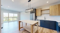 Located at 4 N Hickory Ave in Arlington Heights, IL, this thoughtfully designed single-floor studio offers a contemporary open-concept layout with efficient use of space. The kitchen is a standout feature, showcasing a striking two-tone design with bold navy lower cabinetry and warm natural wood upper cabinets set against a clean white tile backsplash. Stainless steel appliances — including a French door refrigerator, gas range, microwave, and dishwasher — provide a fully equipped culinary space. A wooden kitchen island topped with a white surface sits beneath an industrial-style cage pendant light, adding both function and style. Light-toned hardwood flooring flows seamlessly throughout the unit, lending warmth and continuity to every room. Large floor-to-ceiling sliding glass doors open to a private balcony, inviting abundant natural light and offering open views of the surrounding Arlington Heights neighborhood, including lush tree canopies and the downtown skyline. The bedroom area benefits from expansive windows that further enhance the brightness of the space. The bathroom features gray tile flooring, a full soaking tub with a handheld showerhead, a wood-accented vanity with a white countertop, and clean, neutral wall tones. Recessed lighting and modern black hardware throughout complete the polished, contemporary aesthetic.