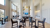 Located in Richmond, TX, this single-floor community clubhouse on Berkeley Knoll Circle offers a thoughtfully designed gathering space defined by dramatic soaring ceilings and an open, airy layout. The main lounge area features striking hexagonal tile flooring, a bold navy sectional sofa, and coordinating accent chairs, all bathed in abundant natural light streaming through floor-to-ceiling glass doors and oversized windows that frame views of the outdoor pool and surrounding grounds.

The social and dining area is anchored by sleek pedestal bistro tables paired with upholstered leather bar-height chairs, arranged beneath a row of statement brass pendant lights. Shiplap accent walls and vertical column detailing lend a clean, modern farmhouse character throughout the space. Built-in open shelving flanks a wall-mounted flat-screen TV, adding both function and visual interest.

A dedicated refreshment station features crisp white cabinetry with matte black hardware, a dark countertop, and a bold geometric-patterned accent wall — ideal for a coffee or beverage bar setup. A restroom with eye-catching patterned tile walls and a vessel sink adds a designer touch. Light wood-tone flooring flows through the primary gathering areas, tying together the space's contemporary, welcoming aesthetic.