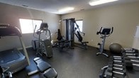 This fitness center offers a practical and functional space designed to support a well-rounded workout routine. The room is outfitted with a selection of cardio equipment, including an elliptical trainer and a recumbent stationary bike, providing options for low-impact cardiovascular exercise. At the center of the space stands a multi-station cable and weight machine, ideal for a range of strength and resistance training exercises. Additional benches are available for stretching or supplementary workouts. The flooring throughout is a durable dark rubber material, engineered to absorb impact and provide stability during exercise. The walls are finished in a neutral beige tone, contributing to a clean and focused environment. Overhead fluorescent lighting ensures the space is well-lit, while multiple windows along the walls allow natural light to filter in, enhancing the overall ambiance. The layout is thoughtfully arranged to allow users to move freely between equipment without congestion. This single-floor fitness facility is straightforward and utilitarian in design, making it an accessible and efficient space for everyday fitness needs.