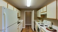 Located at 4401 Beatline Rd. in Long Beach, Mississippi, this single-story space offers a practical and well-organized layout designed for comfortable everyday living. The galley-style kitchen is a standout feature, thoughtfully arranged with white cabinetry lining both walls, providing generous storage space. The kitchen is equipped with a full suite of white appliances, including a refrigerator, electric coil range, and dishwasher, complemented by a double basin sink. Warm hardwood-style flooring runs the length of the kitchen, adding a sense of warmth and continuity to the space. A textured ceiling with overhead fluorescent lighting ensures the kitchen is well-lit and functional. At the far end of the kitchen, a doorway leads to an additional room, suggesting a connected utility or storage area, further enhancing the practicality of the layout. The warm neutral wall tones throughout contribute to a welcoming and cohesive ambiance. Overall, this space presents a straightforward and efficient floor plan ideal for those seeking a functional, low-maintenance living environment in the Long Beach, MS area.