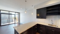This single-floor unit showcases a contemporary design aesthetic defined by clean lines, dark espresso cabinetry, and warm wood-tone flooring throughout the main living areas. The open-concept kitchen serves as the centerpiece of the space, featuring rich dark cabinetry paired with light quartz countertops, a vertically laid white tile backsplash, and stainless steel appliances including a dishwasher, range, microwave, and refrigerator. Globe pendant lights hang above the kitchen peninsula, adding a refined, modern touch. The kitchen flows seamlessly into the living area, which benefits from abundant natural light and direct access to a private balcony through full-height sliding glass doors, framing elevated city views of the surrounding urban skyline.

The bedrooms are carpeted and generously sized, each featuring large floor-to-ceiling windows that capture expansive city and treetop views. The bathroom is finished with large-format light tile flooring, a dark vanity with a stone countertop, a wall-mounted mirror, and a dark tile walk-in shower enclosure. Throughout the unit, a consistent palette of dark, light, and neutral tones creates a cohesive, polished ambiance suited to modern urban living.
