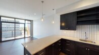 This single-floor unit showcases a contemporary design aesthetic defined by clean lines, dark espresso cabinetry, and warm wood-tone flooring throughout the main living areas. The open-concept kitchen serves as the centerpiece of the space, featuring rich dark cabinetry paired with light quartz countertops, a vertically laid white tile backsplash, and stainless steel appliances including a dishwasher, range, microwave, and refrigerator. Globe pendant lights hang above the kitchen peninsula, adding a refined, modern touch. The kitchen flows seamlessly into the living area, which benefits from abundant natural light and direct access to a private balcony through full-height sliding glass doors, framing elevated city views of the surrounding urban skyline.

The bedrooms are carpeted and generously sized, each featuring large floor-to-ceiling windows that capture expansive city and treetop views. The bathroom is finished with large-format light tile flooring, a dark vanity with a stone countertop, a wall-mounted mirror, and a dark tile walk-in shower enclosure. Throughout the unit, a consistent palette of dark, light, and neutral tones creates a cohesive, polished ambiance suited to modern urban living.