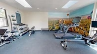 The Mesa del Oso Fitness Center at 5100 Mesa del Oso Rd NE in Albuquerque, New Mexico, is a thoughtfully designed workout facility that packs a comprehensive range of exercise equipment into an efficiently utilized single-room space. The gym features durable rubber flooring throughout, providing a safe and supportive surface for all types of training activities.

Natural light fills the room through two skylights in the textured ceiling, complemented by recessed lighting and additional windows along the walls — including a glass entry door that opens to the outdoors, further brightening the interior. A split-unit climate control system ensures comfort year-round.

The standout visual feature is a sweeping floor-to-ceiling panoramic mural depicting an aerial view of the Albuquerque landscape, complete with colorful hot air balloons — a nod to the region's famous balloon culture and a striking focal point that energizes the space.

Equipment includes an Echelon rowing machine, a Nautilus treadmill, an elliptical trainer, and an Echelon connected fitness bike for cardio training. Strength training is well-supported with a Hoist functional trainer cable machine, a dumbbell rack with a full range of weights, medicine balls, a weight bench, and a dedicated free weight area. Informational fitness posters and clearly posted facility rules round out the practical, user-focused environment.