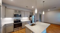 This thoughtfully designed single-floor residence offers a seamless open-concept layout that connects the living and kitchen areas, creating a bright and functional living environment. The kitchen is a standout feature, showcasing grey shaker-style cabinetry with glass-front upper panels, white quartz countertops, a classic subway tile backsplash, and a full suite of stainless steel appliances including a range, microwave, and refrigerator. A central island with an undermount sink and elegant pendant lighting adds both style and utility. The expansive living area is finished with warm hardwood-style flooring, a ceiling fan, and access to a private balcony through glass-panel doors, allowing natural light to flow throughout. The primary bathroom features a wide dual-sink vanity with white countertops, grey cabinetry, a large mirror with vanity lighting, and tile flooring. A separate walk-in shower is enclosed by a frameless sliding glass door. The generous walk-in closet is equipped with wire shelving on multiple walls, offering abundant storage. A dedicated in-unit laundry space houses a full-size washer and dryer with additional wire shelving above. Throughout the residence, grey-toned interior doors and clean, neutral finishes contribute to a cohesive and contemporary aesthetic.