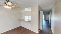 This single-floor space showcases a practical and open layout designed to maximize functionality. The kitchen area is positioned along one wall and features white cabinetry, a tile countertop with a built-in sink, and a pass-through window that connects to the adjacent living area. This pass-through design creates a sense of openness between the spaces while maintaining a defined kitchen zone. Under-cabinet lighting adds a functional element above the workspace.

The flooring throughout is a warm-toned hardwood-style material that runs continuously from the kitchen area into the living space, providing visual cohesion across the floor plan. The walls are finished in a neutral white, complementing the light overall palette of the space.

A louvered bi-fold door is visible near the transition between the kitchen and the living area, likely concealing a storage closet or utility space. Beyond it, the living area benefits from natural light streaming in through what appears to be a large window or sliding glass door. The textured ceiling is consistent throughout, adding a classic finishing detail to the interior.